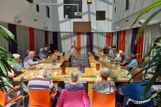 Een grote groep mensen in een zaal, aan tafel in theater opstelling, kijkend naar een presentatie